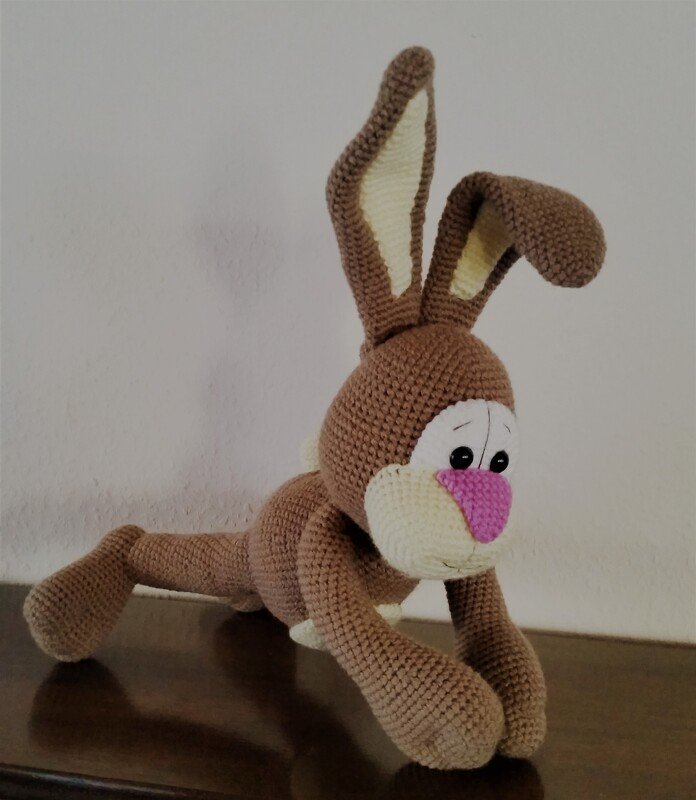 Braunes Amigurumi-Häschen mit rosa Nase und weißen Gesichtspartien, kauernd auf dunkler Oberfläche