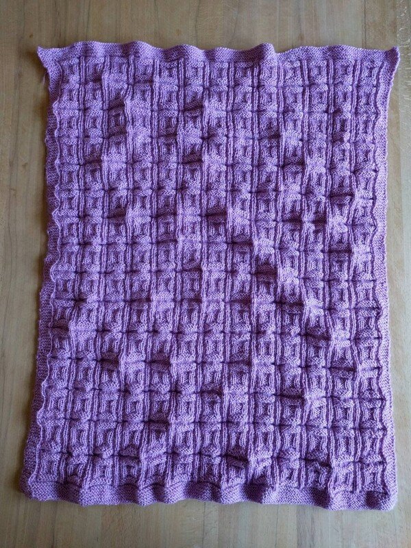 Pastelllila gestrickte Babydecke mit Relief-Karomuster und kraus gestricktem Rand auf Holzboden