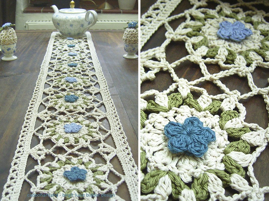 Nahaufnahme eines gehäkelten Tischläufers im Granny-Stil mit cremefarbenen, grünen und blauen Blütenmotiven, der auf einem Holztisch liegt.