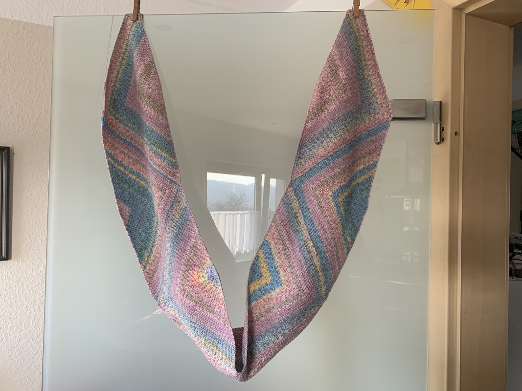 Gehäkeltes Dreieckstuch in Pastellrosa, Blau und Gelb, dekorativ aufgehängt vor hellem Hintergrund.