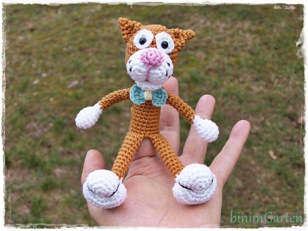 Gehäkelte braune Amigurumi-Katze mit weißen Pfoten und hellblauer Fliege, in einer Hand vor unscharfem Gras.