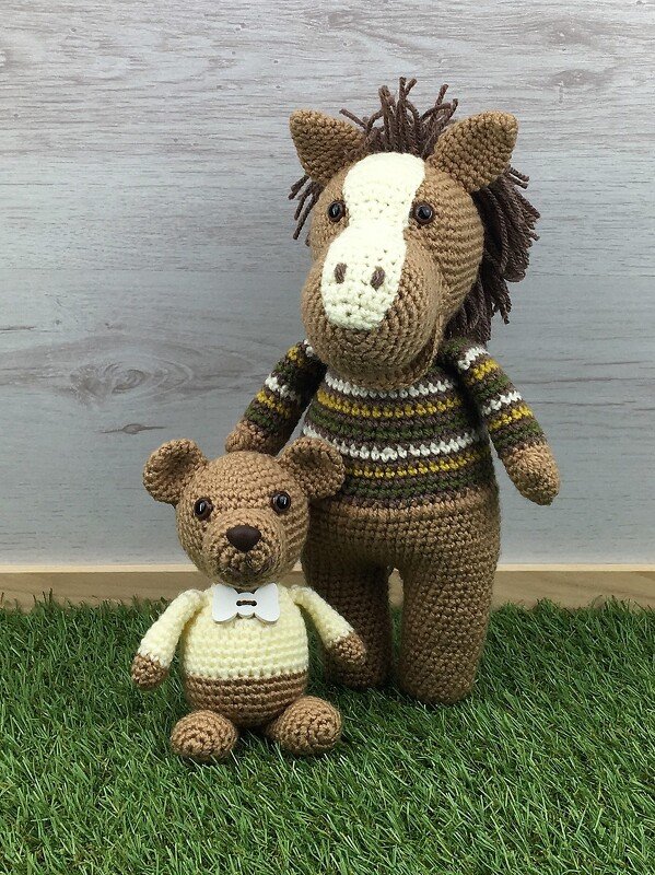 Gehäkeltes Amigurumi-Pferd im Streifenpullover hinter einem sitzenden Bären mit Fliege vor Holzhintergrund auf grünem Rasenteppich.