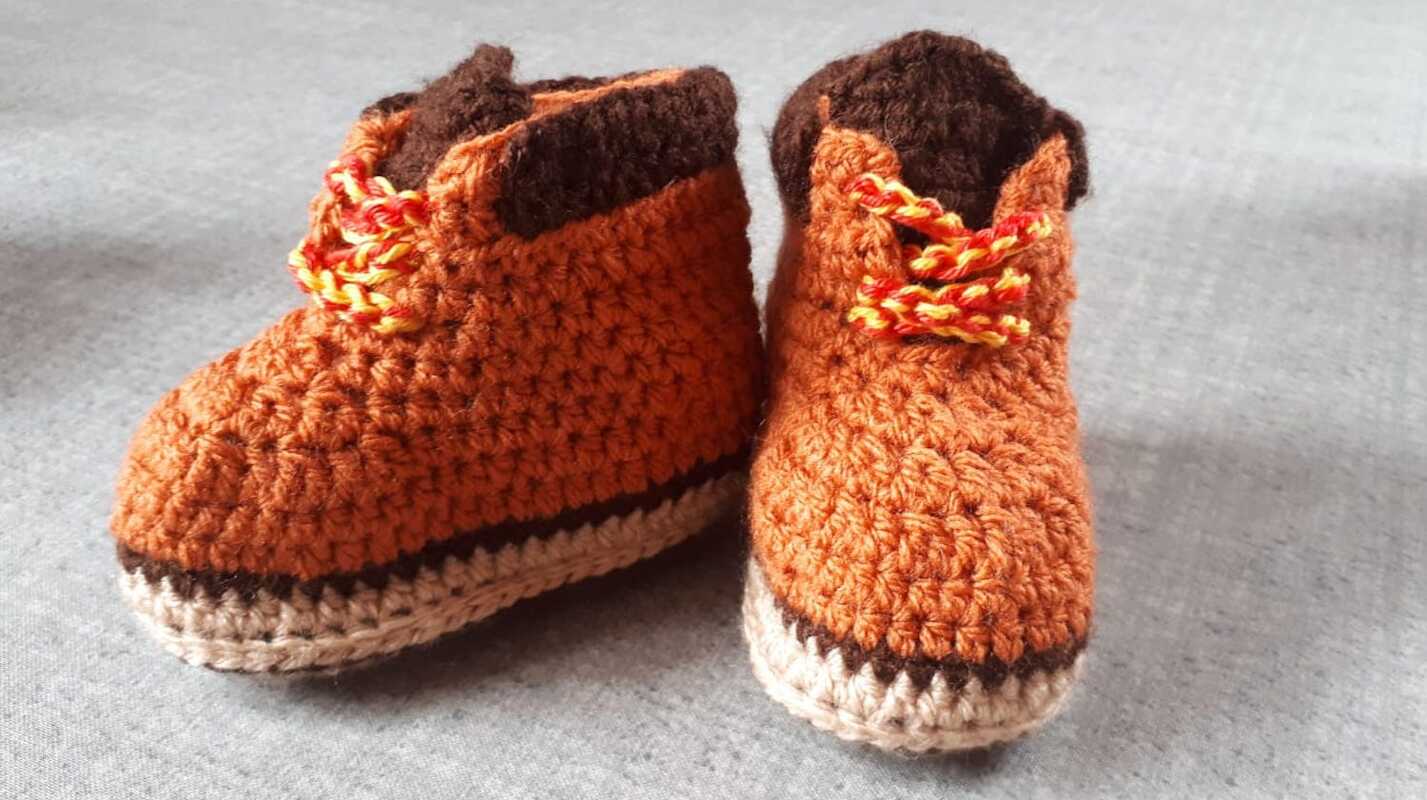 Orangene gehäkelte Babyschuhe mit braunem Rand und gelb-roter Schnürung auf grauem Stoff