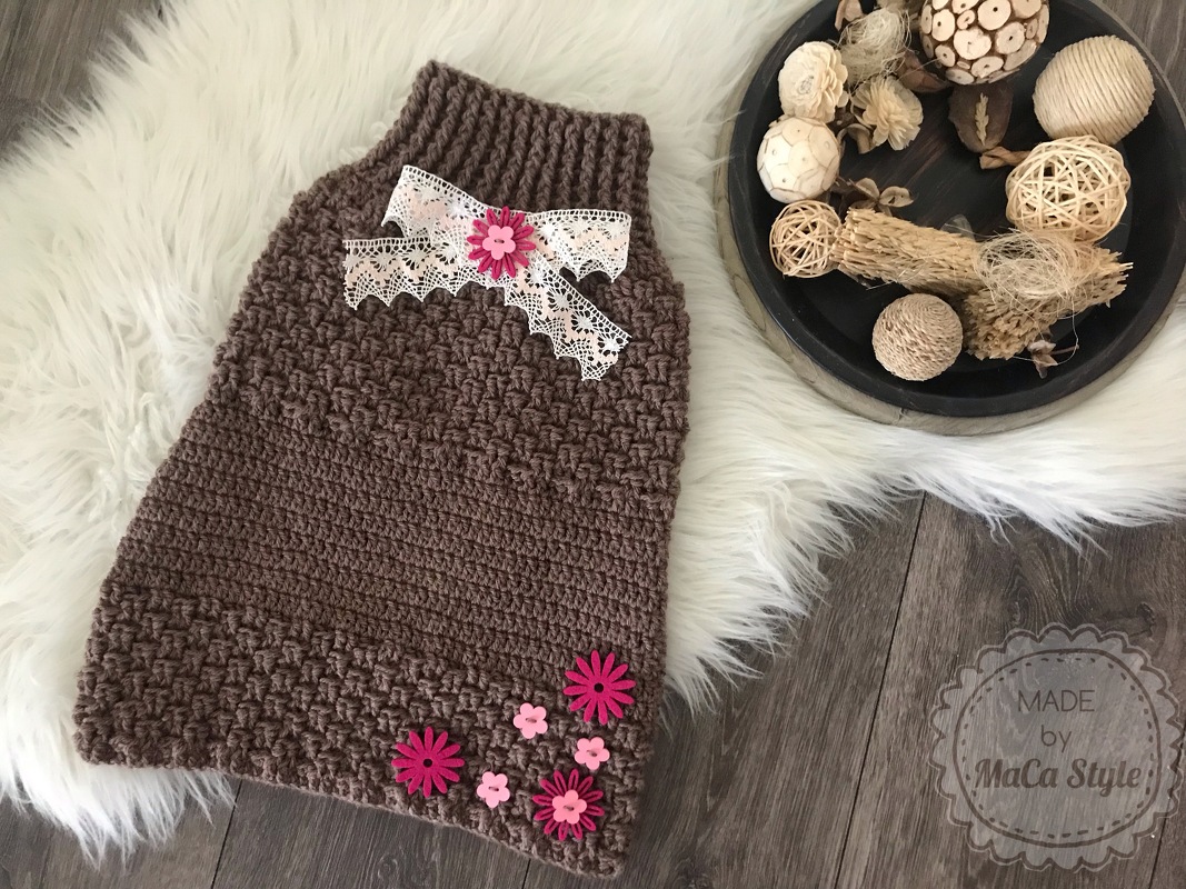 Brauner gehäkelter Babyschlafsack mit weißer Spitzenschleife und pinken Filzblüten auf weißem Felluntergrund, neben Naturdekoration.