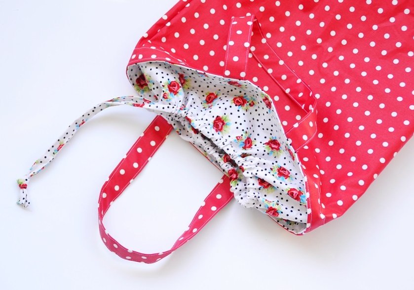 Rote Stofftasche mit weißen Punkten, Tragehenkeln und gerafftem Tunnelzug, aus dem ein geblümtes Innenfutter hervorschaut