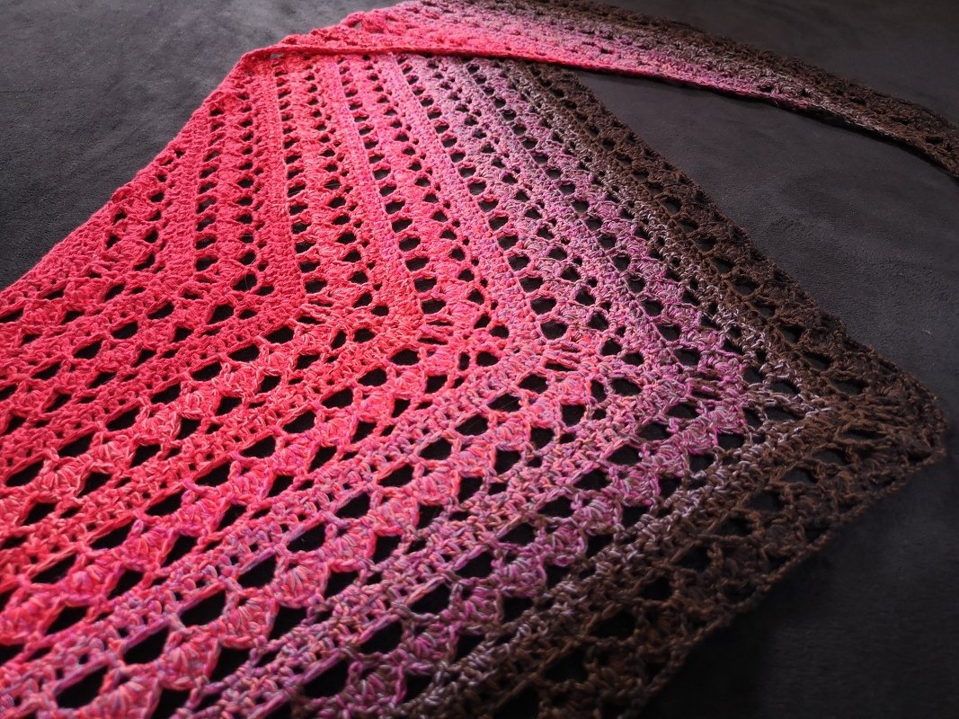 Gehäkeltes Dreieckstuch mit Farbverlauf von Pink über Violett zu Braun auf dunklem Hintergrund.