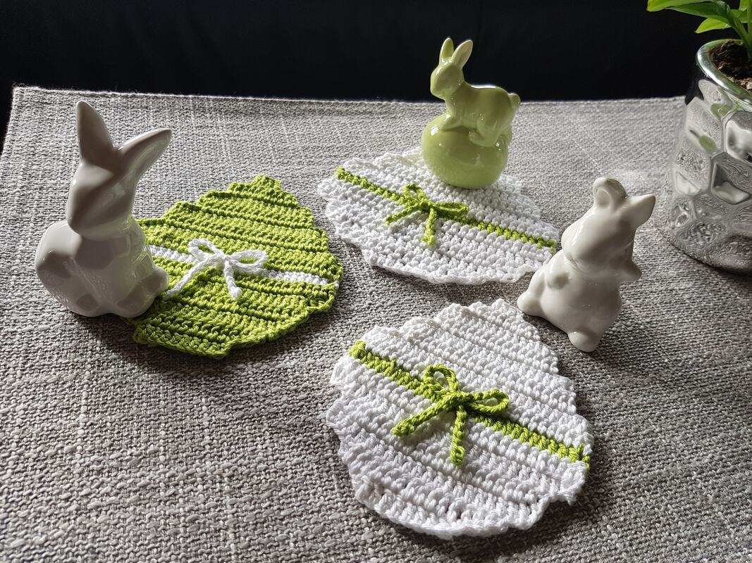 Drei gehäkelte Osterei‑Untersetzer in Grün und Weiß mit Schleifen und Hasenfiguren auf grauem Tischtuch.