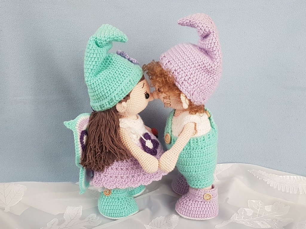 Zwei gehäkelte Amigurumi-Puppen in Mint und Lila mit Zipfelmützen und Flügeln, die sich an den Händen halten
