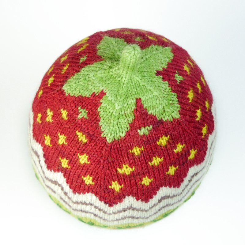 Red knitted strawberry hat with yellow seed details, green leaf top and cream wavy brim.