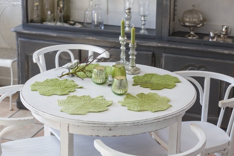 Weißer Tisch mit hellgrünen gestrickten Blatt-Deckchen, silbernen Kerzenhaltern und grünen Glaswindlichtern