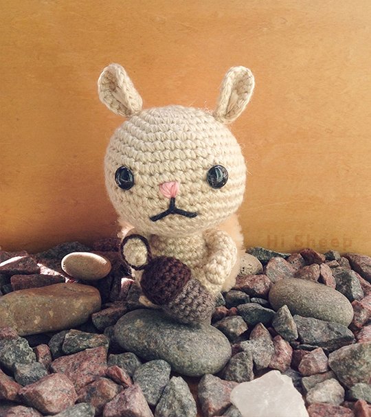 Beige crocheted amigurumi bunny with dark round eyes and pink nose holding a brown crocheted acorn, seated on gray and reddish rocks.