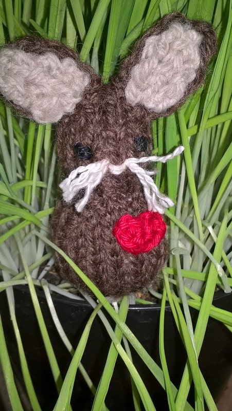 Kleiner brauner Amigurumi-Osterhase mit rotem Herz zwischen grünen Grashalmen