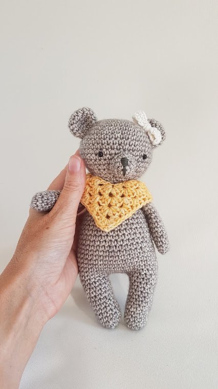 Small grey crocheted teddy bear wearing a yellow bandana and a white bow, held by a hand against a plain light background.