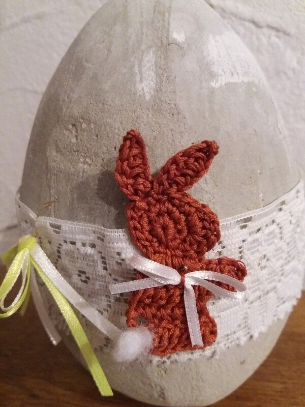 Gehäkelter rotbrauner Hase mit weißem Bommelschwanz und Schleife auf grauem Dekoei mit weißer Spitze und Bändern.