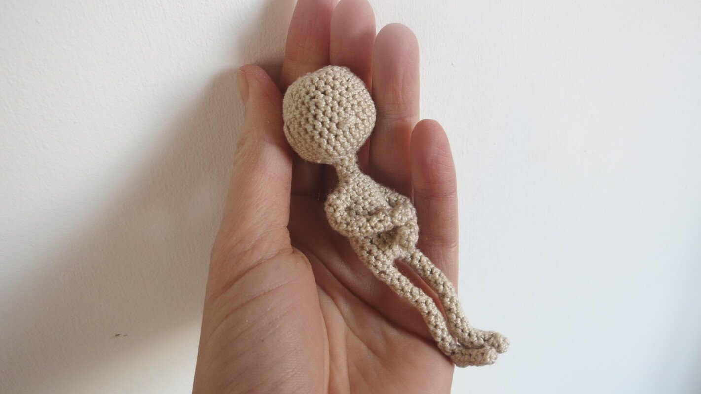 Hand holding a small beige crocheted amigurumi doll body with round head and long legs
