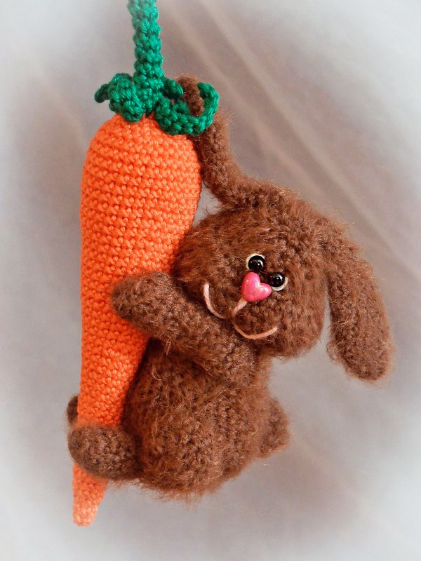 Gehäkelter brauner Amigurumi-Hase mit rosa herzförmiger Nase hält eine orangefarbene Karotte mit grünen Blättern.