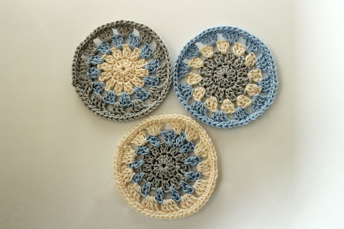 Drei runde, handgehäkelte Untersetzer in Creme, Hellblau und Grau mit sternförmigem Muster auf hellem Hintergrund