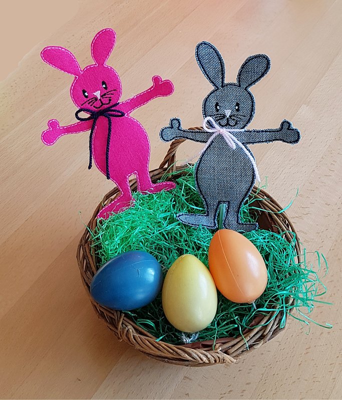 Zwei gestickte Hasenfiguren in Pink und Grau im Weidenkörbchen mit grünem Ostergras und drei bunten Ostereiern.