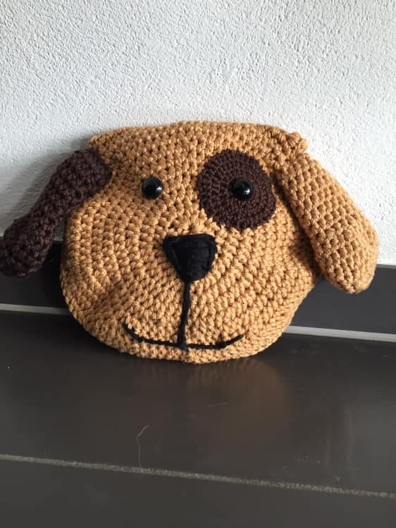 Rundes gehäkeltes Kissen in Form eines Hundegesichts mit braunem Fleck, Schlappohren und schwarzer Nase.