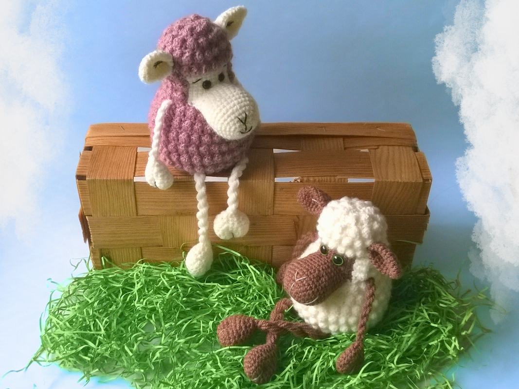 Zwei kuschelige gehäkelte Amigurumi‑Schafe in Lila und Weiß mit Korb und grünem Dekogras vor blauem Himmel.