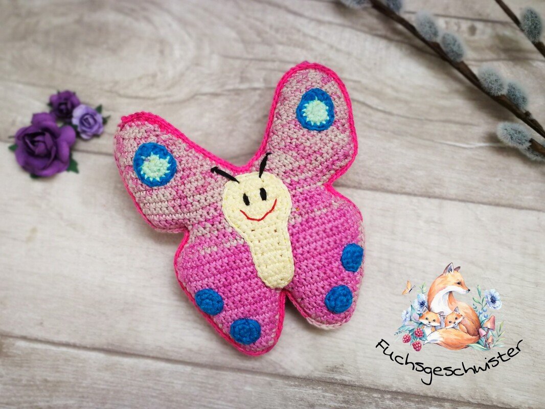 Gehäkelter rosa Schmetterling mit lächelndem Gesicht und blauen Punkten auf hellem Holzuntergrund