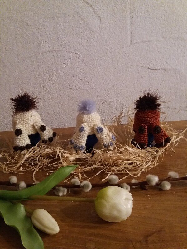 Drei gehäkelte Eierwärmer in Cremeweiß, Blau und Rostrot mit Pompons in Strohbett, neben Weidenkätzchen und Tulpe auf Holz.