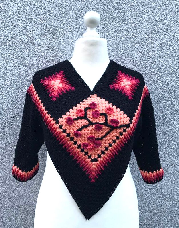 Schwarz-rosa gehäkelter Damenponcho mit Granny-Dreieck und Blumenapplikation auf Schaufensterpuppe vor grauer Wand