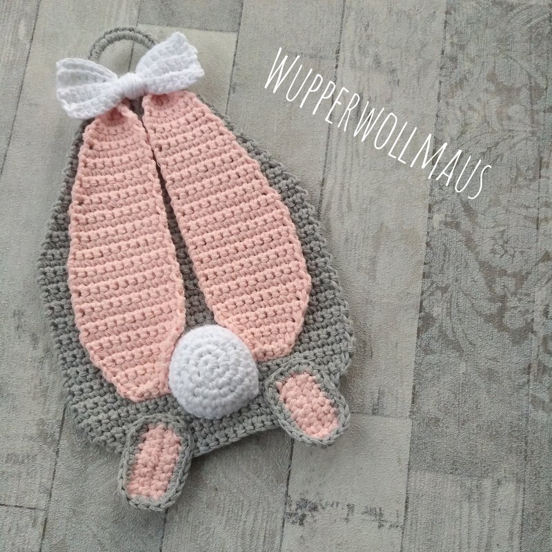 Gehäkelter Hasen-Topflappen in Grau mit rosa Ohren, weißer Schleife und Bommel auf hellem Holzhintergrund.