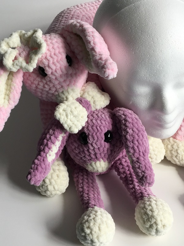 Zwei gehäkelte Plüschhasen in Rosa und Lila mit cremefarbenen Akzenten neben einem Styroporkopf