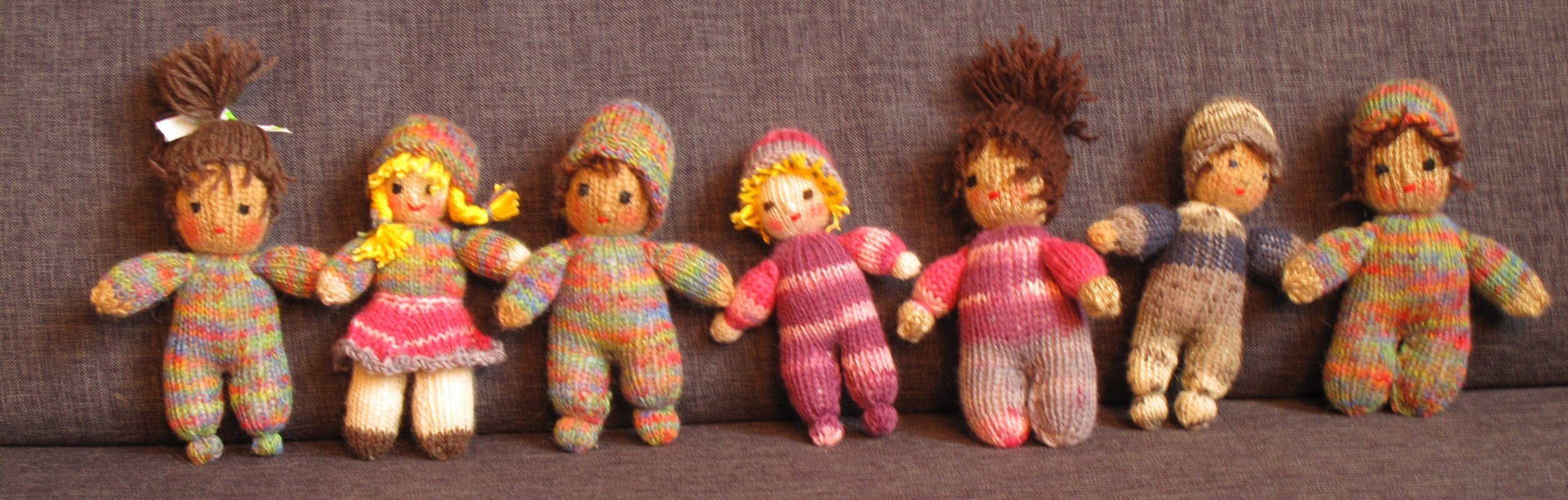 Sieben gestrickte Amigurumi‑Puppen in bunten Garnen nebeneinander auf grauem Stoff