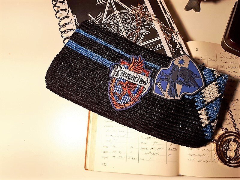 Schwarze gehäkelte Tasche mit blauen Streifen und Wappen-Patches auf offenem Buch
