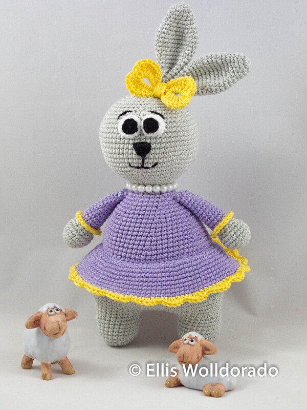 Gehäkelter grauer Amigurumi-Hase im lila Kleid mit gelber Schleife und zwei kleinen Schafen
