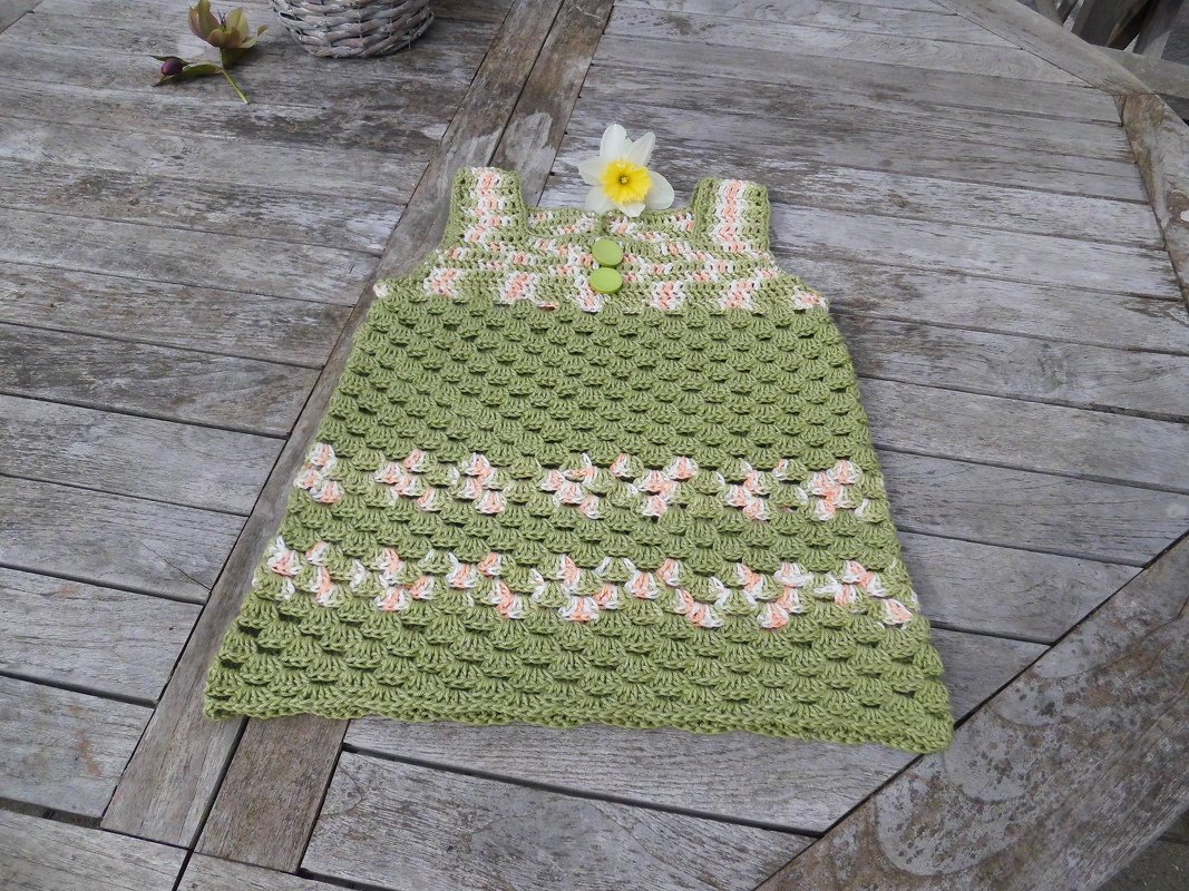 Gehäkeltes hellgrünes Babykleid mit Muschelmuster und Zartrosa‑Weiß‑Akzenten, liegend auf einem Holztisch