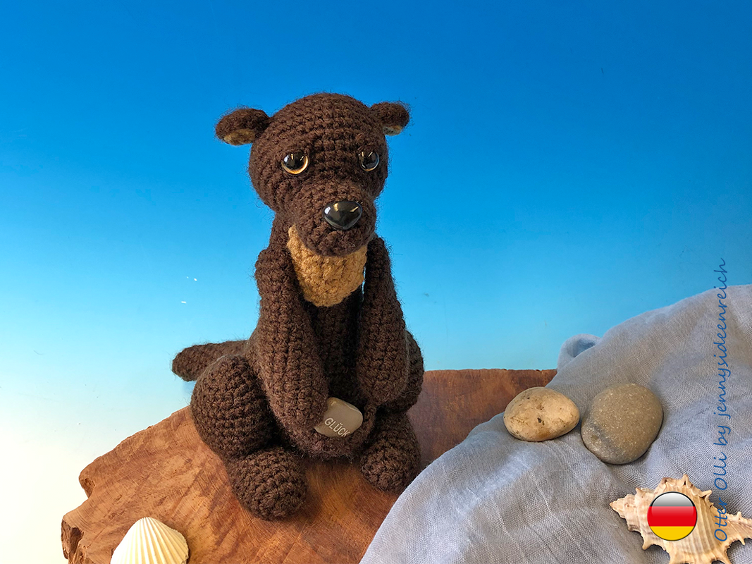 Brauner Amigurumi-Otter mit hellem Brustteil hält Stein „Glück“, sitzt auf Holzscheibe neben Tuch, Steinen und Muscheln
