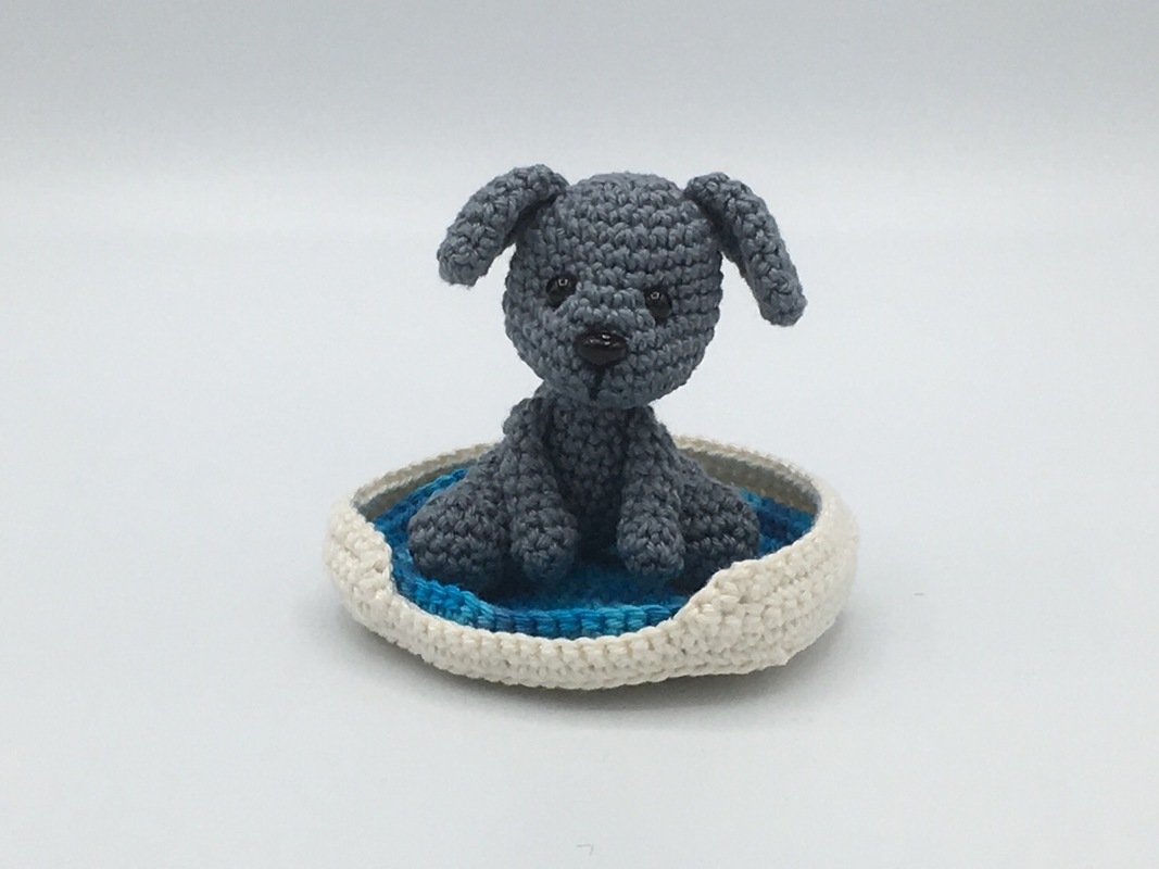 Gehäkelter grauer Amigurumi‑Hund im runden weiß‑blauen Körbchen