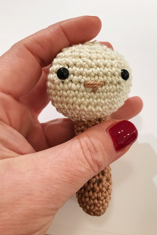 Hand hält kleine gehäkelte Babyrassel mit cremefarbenem Kopf und braunem Griff