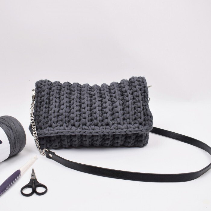 Dunkelgraue gehäkelte Clutch aus dickem Textilgarn mit Metallkette, schwarzem Lederriemen, daneben Garn, Häkelnadel und Schere auf weißem Hintergrund