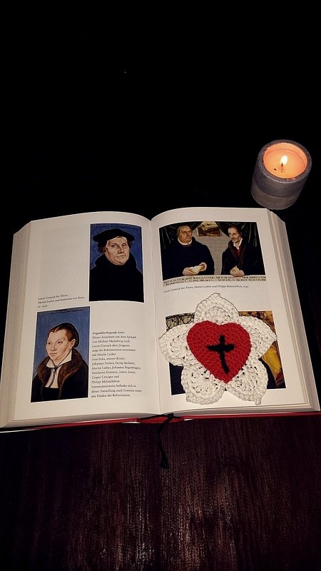 Weiße gehäkelte Blumenapplikation mit rotem Herz und schwarzem Kreuz auf einem aufgeschlagenen Buch, daneben eine brennende Kerze.