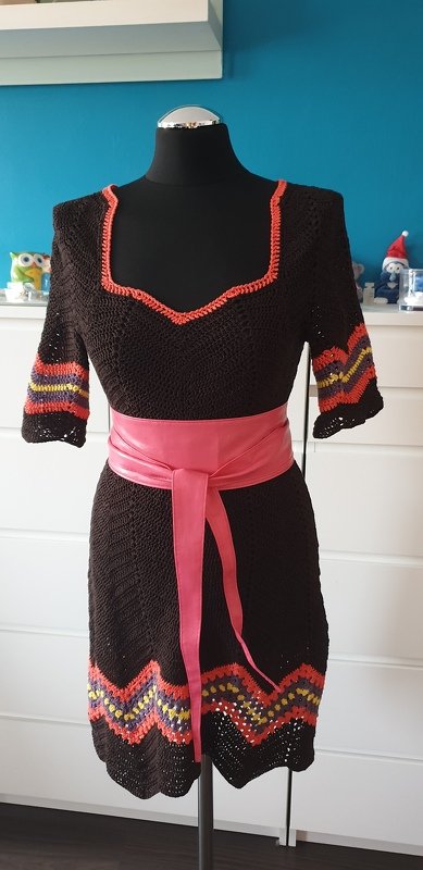 Dunkelbraunes Häkelkleid mit korallenfarbener Einfassung, bunten Zickzackstreifen an Ärmeln und Saum sowie rosa Taillengürtel auf einer schwarzen Schaufensterpuppe.