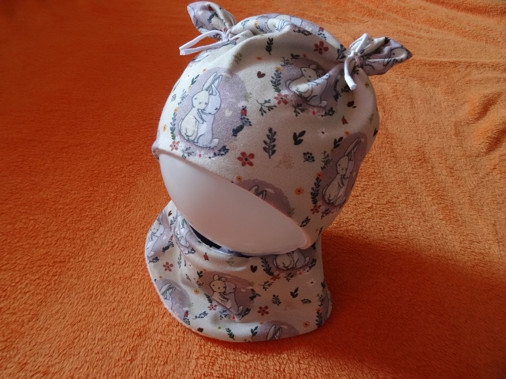 Baby-Mütze und passendes Halstuch mit Hasen- und Blumenmuster auf orangefarbenem Hintergrund.