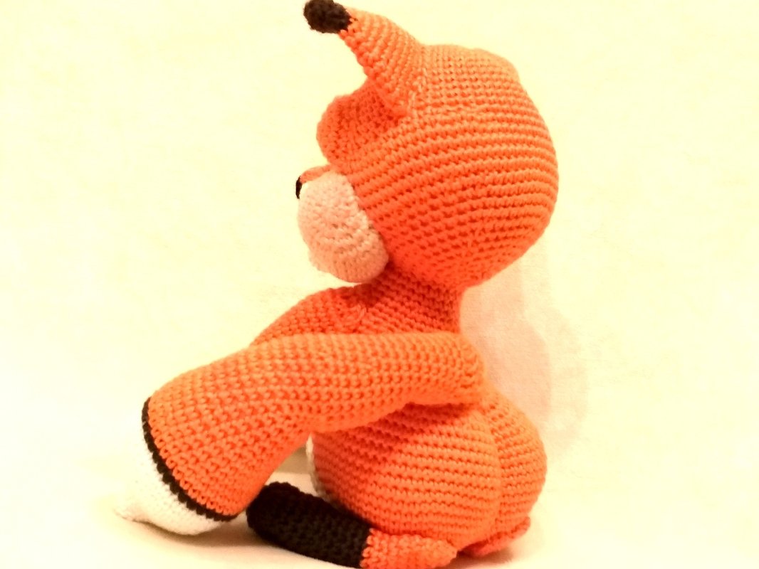 Side view of an orange crocheted amigurumi fox with white muzzle and black accents on a plain light background.