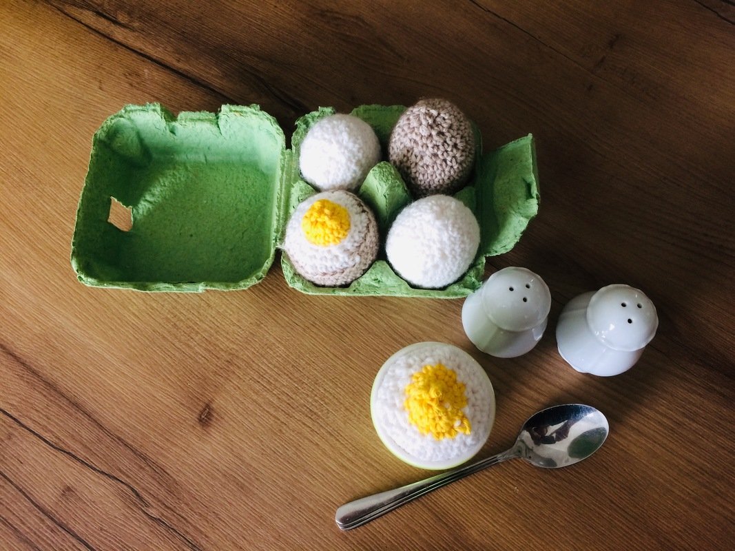 Gehäkelte Eier, teils als Spiegelei, im grünen Eierkarton und im Eierbecher mit Löffel und Salzstreuern auf Holztisch.