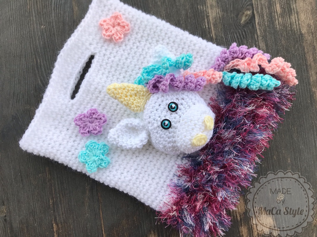 Weiße gehäkelte Tasche mit Einhornkopf, pastellfarbener Mähne, Blumenapplikationen und buntem Fransenrand auf dunklem Holz