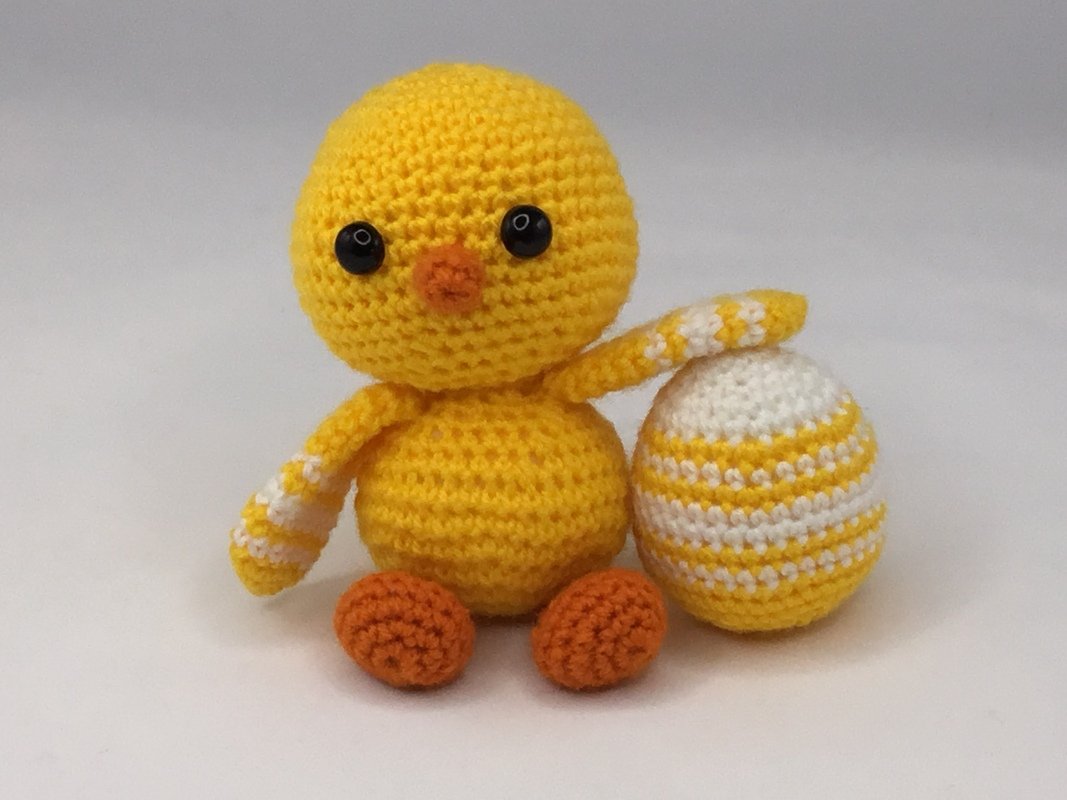 Gelbes gehäkeltes Amigurumi-Küken mit orangefarbenen Füßen neben einem gelb-weiß gestreiften Ei auf hellem Hintergrund.