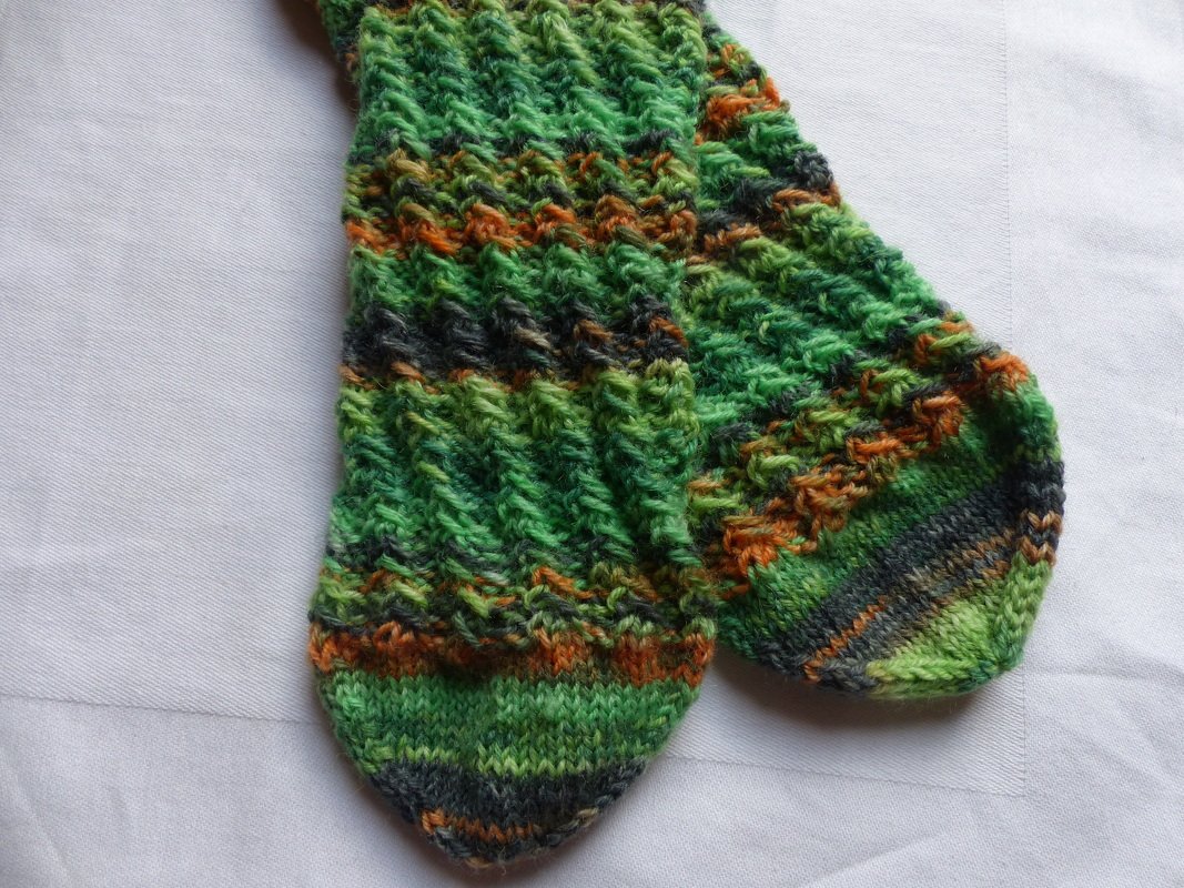Gestricktes Paar Socken in verschiedenen Grüntönen mit orange-grauen Streifen und strukturiertem Muster, auf hellem Stoff liegend