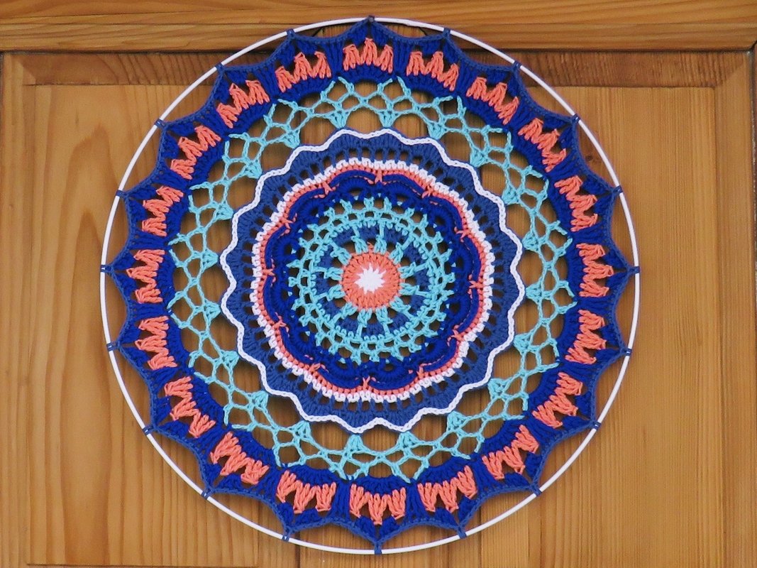 Rundes gehäkeltes Mandala in Blau-, Türkis- und Koralltönen auf weißem Ring vor Holztür