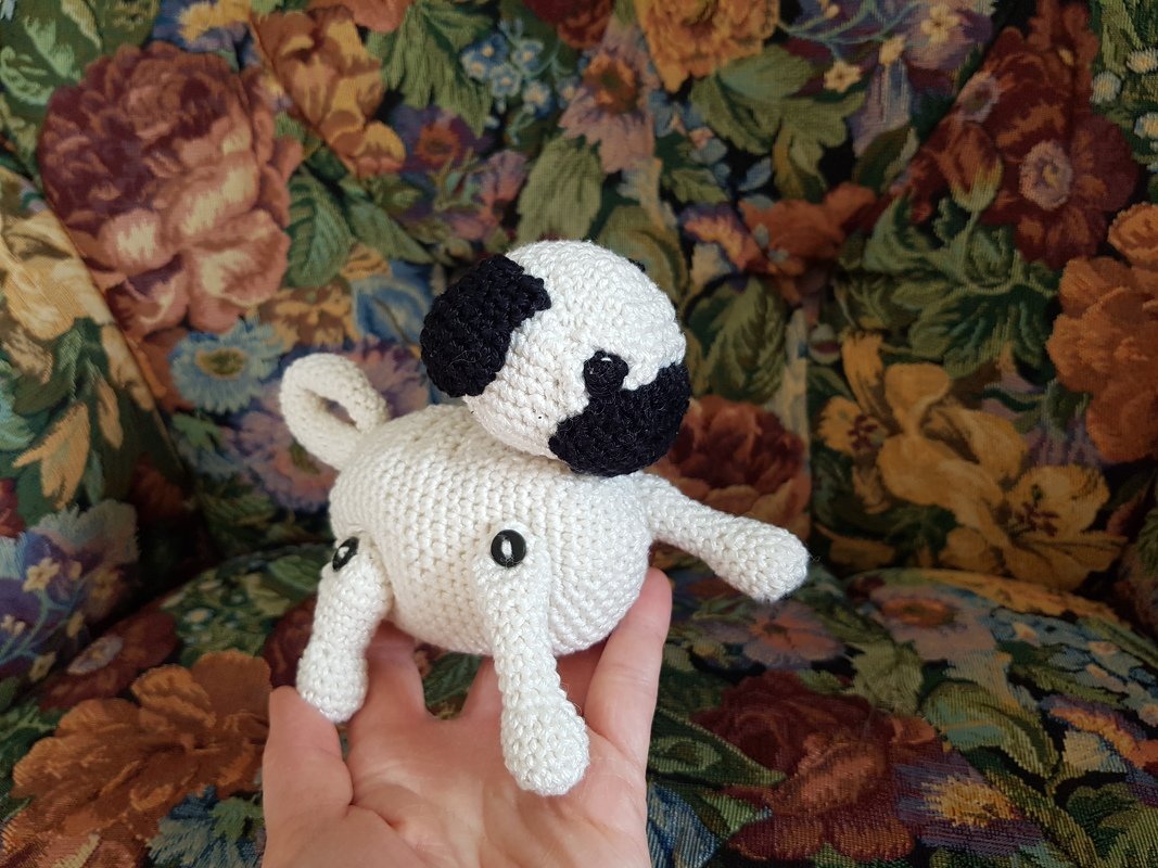 Gehäkelter weißer Amigurumi-Hund mit schwarzen Flecken, gehalten in einer Hand vor gemustertem Polster