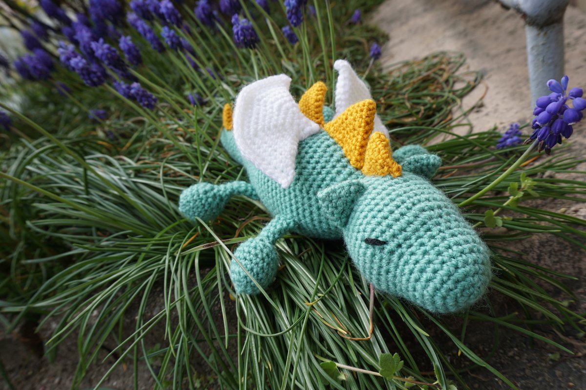 Türkiser Amigurumi-Drache mit weißen Flügeln und gelben Stacheln liegt im Gras neben violetten Blüten.
