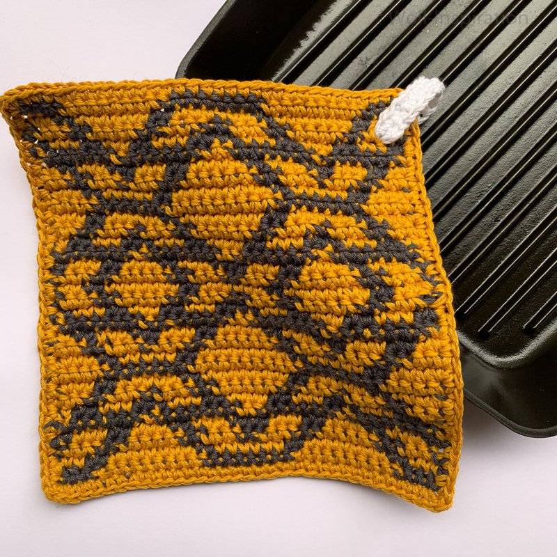 Quadratischer gehäkelter Topflappen in Senfgelb und Dunkelgrau mit grafischem Muster und weißer Aufhängeschlaufe, teilweise auf einer schwarzen Grillpfanne.