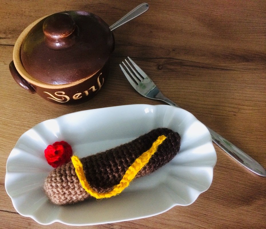 Gehäkelte Wurst mit gelbem und rotem Garn in weißer Schale, Keramik-Senftopf und Gabel auf Holztisch.